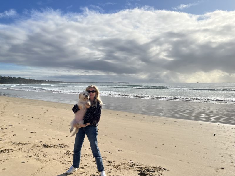 Woman standing on a sandy beach with her dog Eliza, enjoying a sunny day by the ocean.
