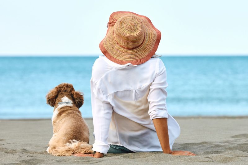 Woman in a wide-brim hat sitting on the sand with her small dog, both facing the ocean at Surfers Paradise on the Gold Coast—relaxed dog-friendly holiday scene.